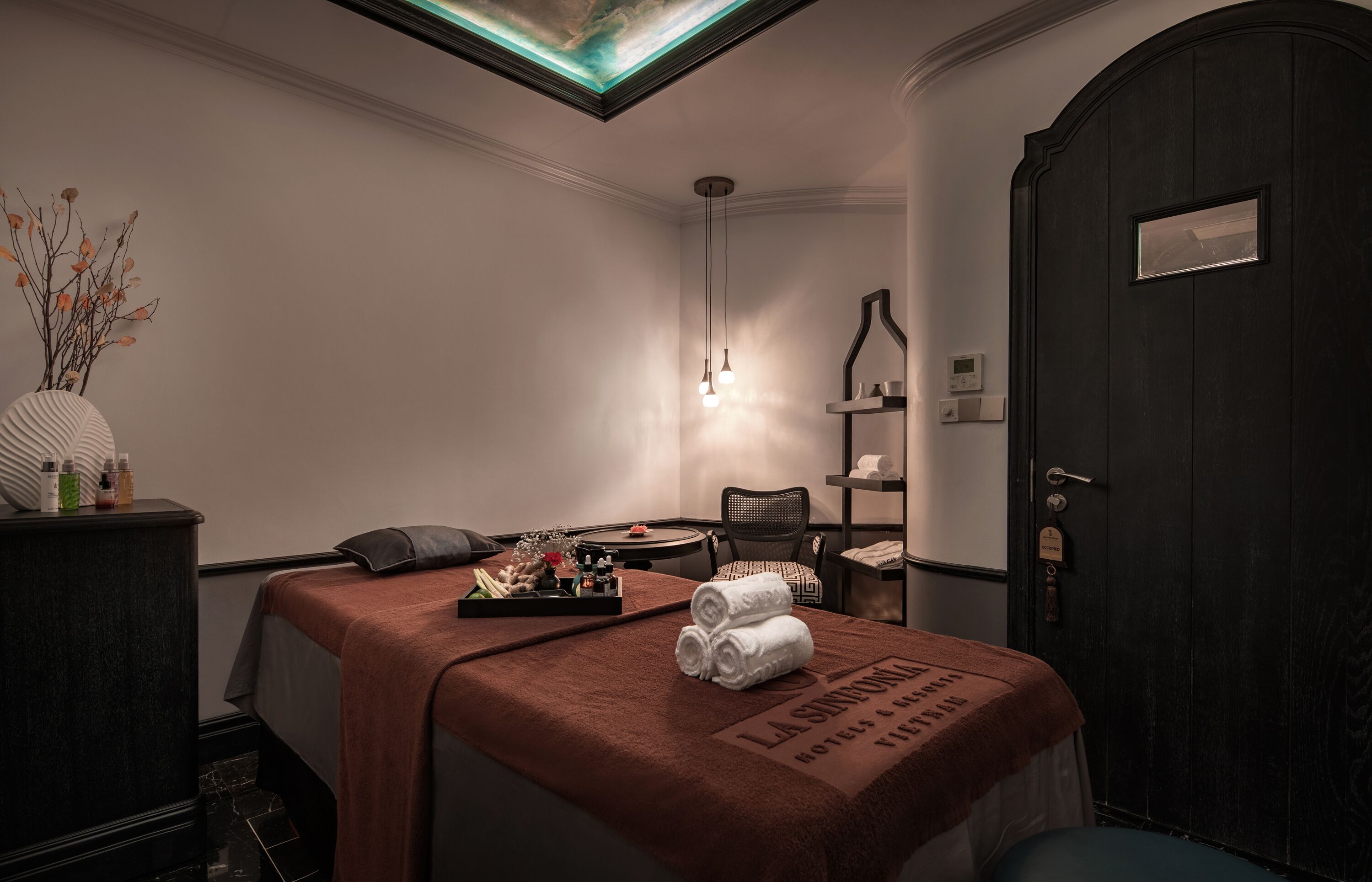 Hotelkamer met een massagetafel, een donker houten deur, een klein plankje met toiletartikelen en een vaas met gedroogde takken.