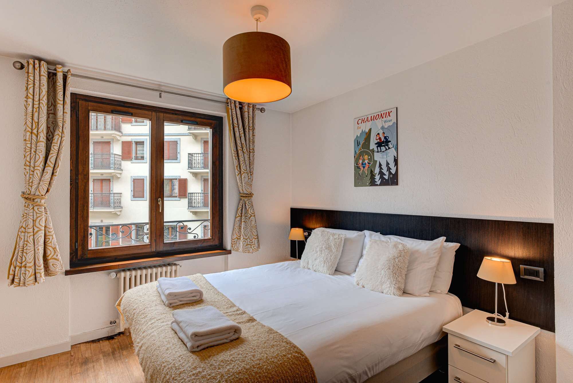 Hotellihuone, jossa on suuri sänky, yöpöytä, lamppu, ikkuna verhoilla ja juliste seinällä.