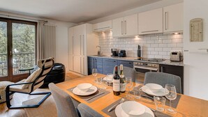 A modern kitchen with white cabinets, a dining table set for four, and a view of the outdoors through large windows. - Apartment Cosmiques - Alpes Agency - Central Chamonix - Sleeps 4 (Chamonix-Mont-Blanc)