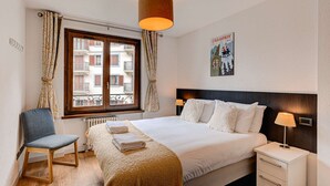 A hotel room with a bed, a chair, a nightstand, a lamp, a window with curtains, and a poster on the wall. - Apartment Cosmiques - Alpes Agency - Central Chamonix - Sleeps 4 (Chamonix-Mont-Blanc)