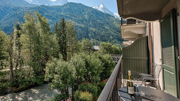 A balcony with a table and chairs overlooking a forested valley and mountains. - Apartment Cosmiques - Alpes Agency - Central Chamonix - Sleeps 4 (Chamonix-Mont-Blanc)