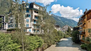 A mountain town with multi-story buildings, a narrow river, and lush greenery. - Apartment Cosmiques - Alpes Agency - Central Chamonix - Sleeps 4 (Chamonix-Mont-Blanc)