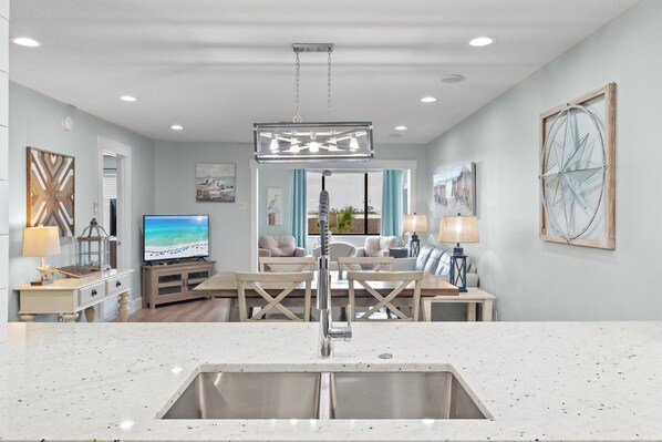 A modern kitchen with a stainless steel sink, a dining area with a wooden table and chairs, and a living room with a television and artwork on the walls.
