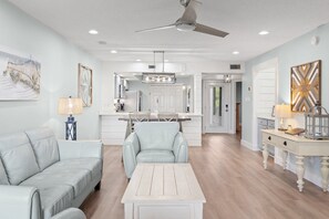 A modern living room with a sofa, a coffee table, and a dining area with chairs and a kitchen in the background. - "Salty Sun" - Golf Cart (included), Tennis, Pools, Golf, Beach... it's all here! (Destin)
