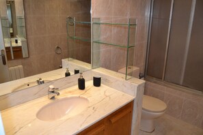 Un baño moderno con lavamanos de mármol, repisas de vidrio, un inodoro y una cabina de ducha de vidrio.