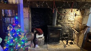 A stone fireplace with a wood-burning stove, a Christmas tree with colorful lights, and a rustic decor including a lantern and a clock. - COTSWOLD HISTORIC COTTAGE, WITH LARGE GARDEN (Naunton)