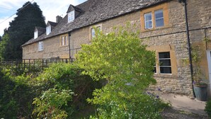 A stone building with multiple windows and a small garden area with a tree and flowers. - COTSWOLD HISTORIC COTTAGE, WITH LARGE GARDEN (Naunton)