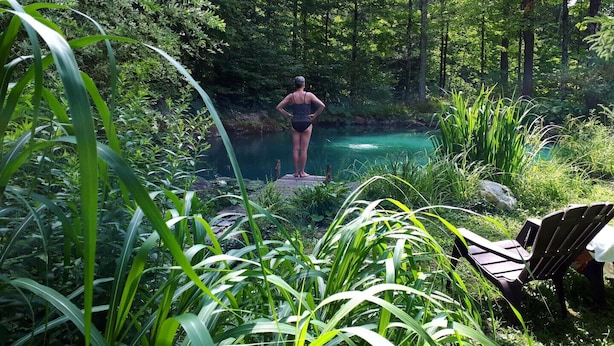 Une personne se tient sur une jetée en bois, surplombant un étang aux eaux bleues et limpides, entouré d’une végétation luxuriante.