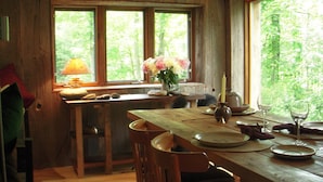 Un coin repas rustique en bois, avec une table dressée pour deux personnes, une fenêtre donnant sur un paysage verdoyant et une lampe posée sur le rebord de la fenêtre.