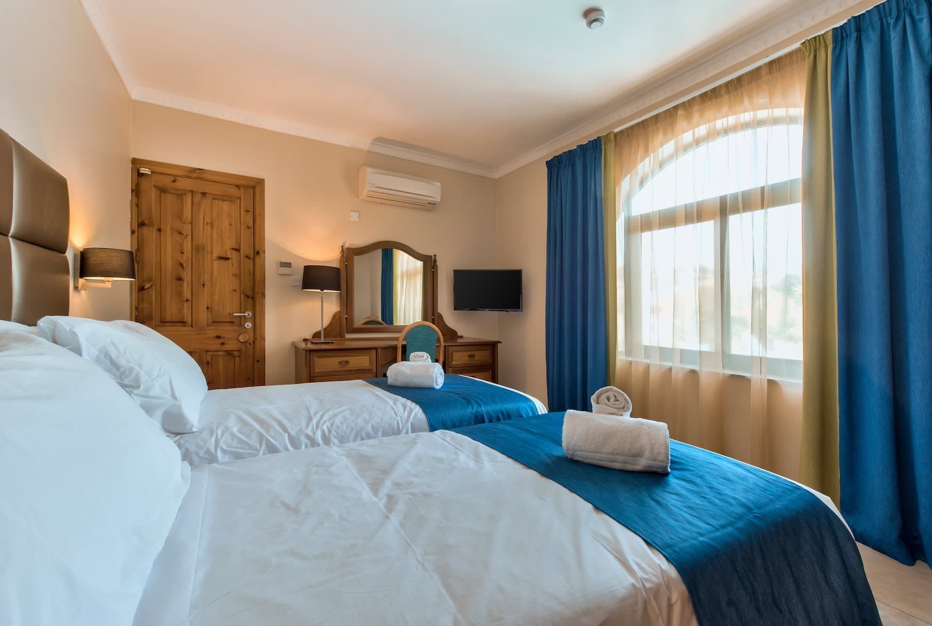 A hotel room with two beds, a wooden wardrobe, a desk with a mirror, a flat-screen TV, and a large window with blue curtains.