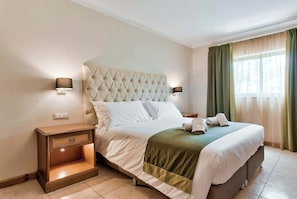 A hotel room with a tufted headboard, a wooden nightstand with drawers, a bed with white linens and a green throw, and a window with sheer curtains.