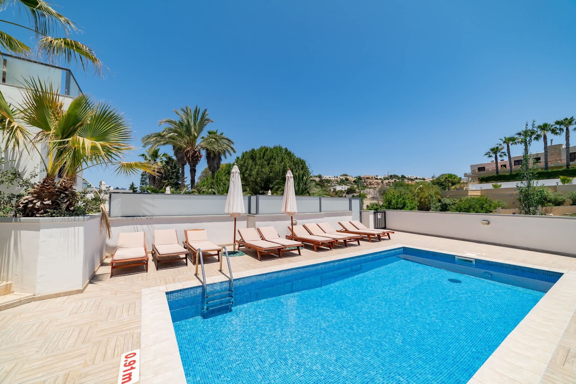 Une piscine rectangulaire à l’eau d’un bleu limpide, entourée de chaises longues et de parasols, avec des palmiers et un ciel dégagé en arrière-plan.