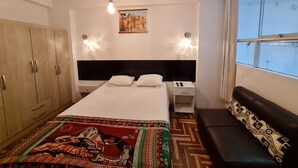 Hotellihuone, jossa on sänky, vaatekaappi, yöpöydät, penkki ja suuri ikkuna.