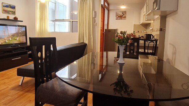 Moderni olohuone, jossa on lasinen ruokapöytä, televisio sekä keittiöalue, jossa on kaappeja ja liesi.