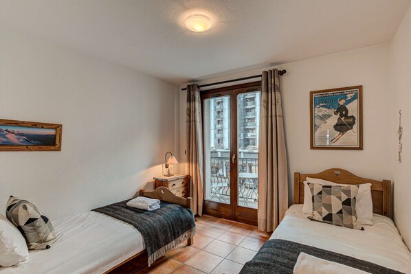 A hotel room with two beds, a window with a view of buildings, a painting on the wall, and a poster. - Apartment Kitson - Alpes Agency - Central Chamonix - Sleeps 6 (Chamonix-Mont-Blanc)