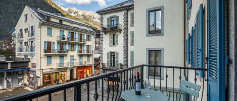 A balcony with a table and chairs, overlooking a street with buildings and a mountain in the background.