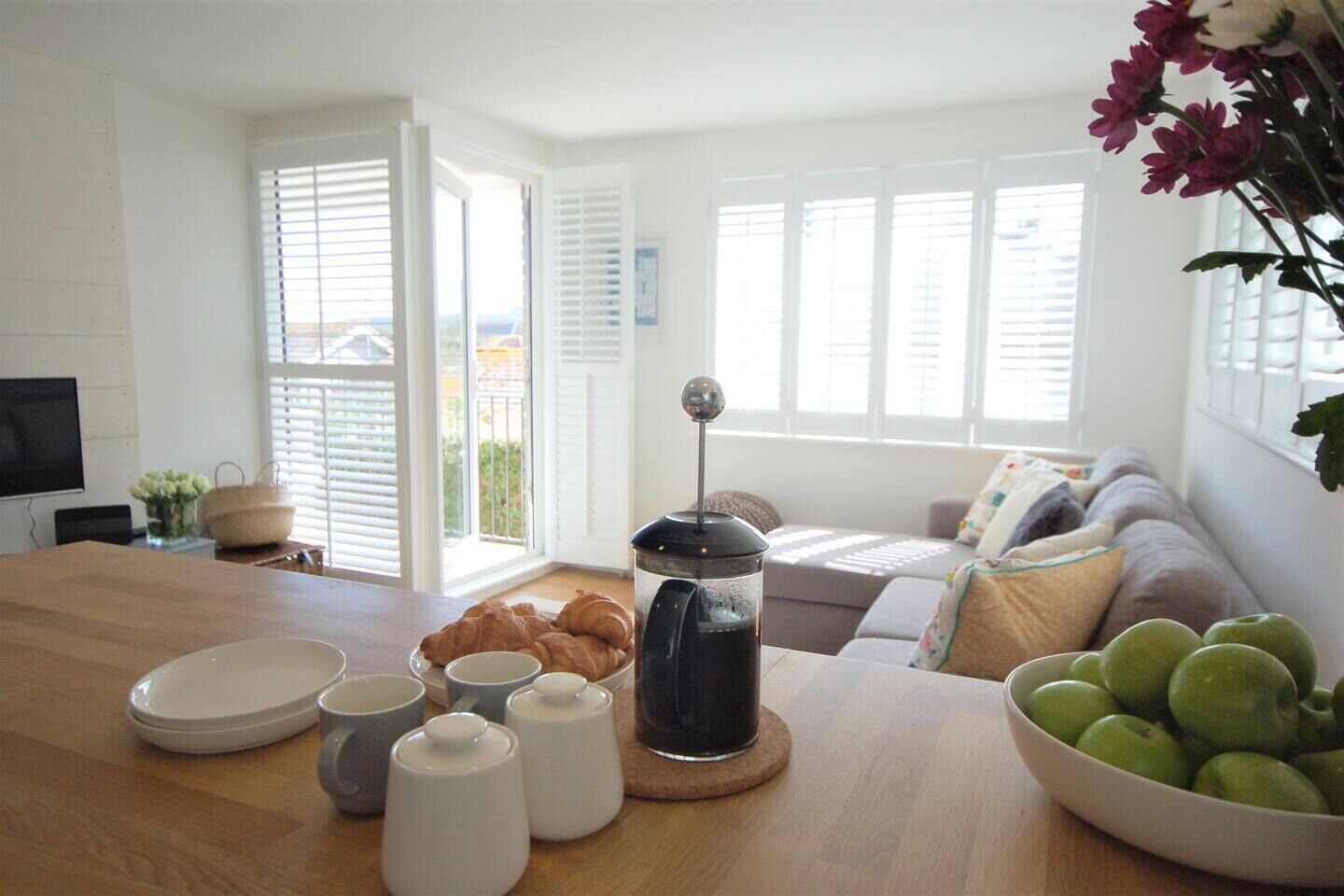 A bright living room with a wooden table, a coffee maker, croissants, and a bowl of apples.