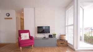 Una sala de estar con un sillón rosa, un mueble gris para equipo audiovisual con televisor, un tapete blanco y vistas amplias a través de ventanas grandes.