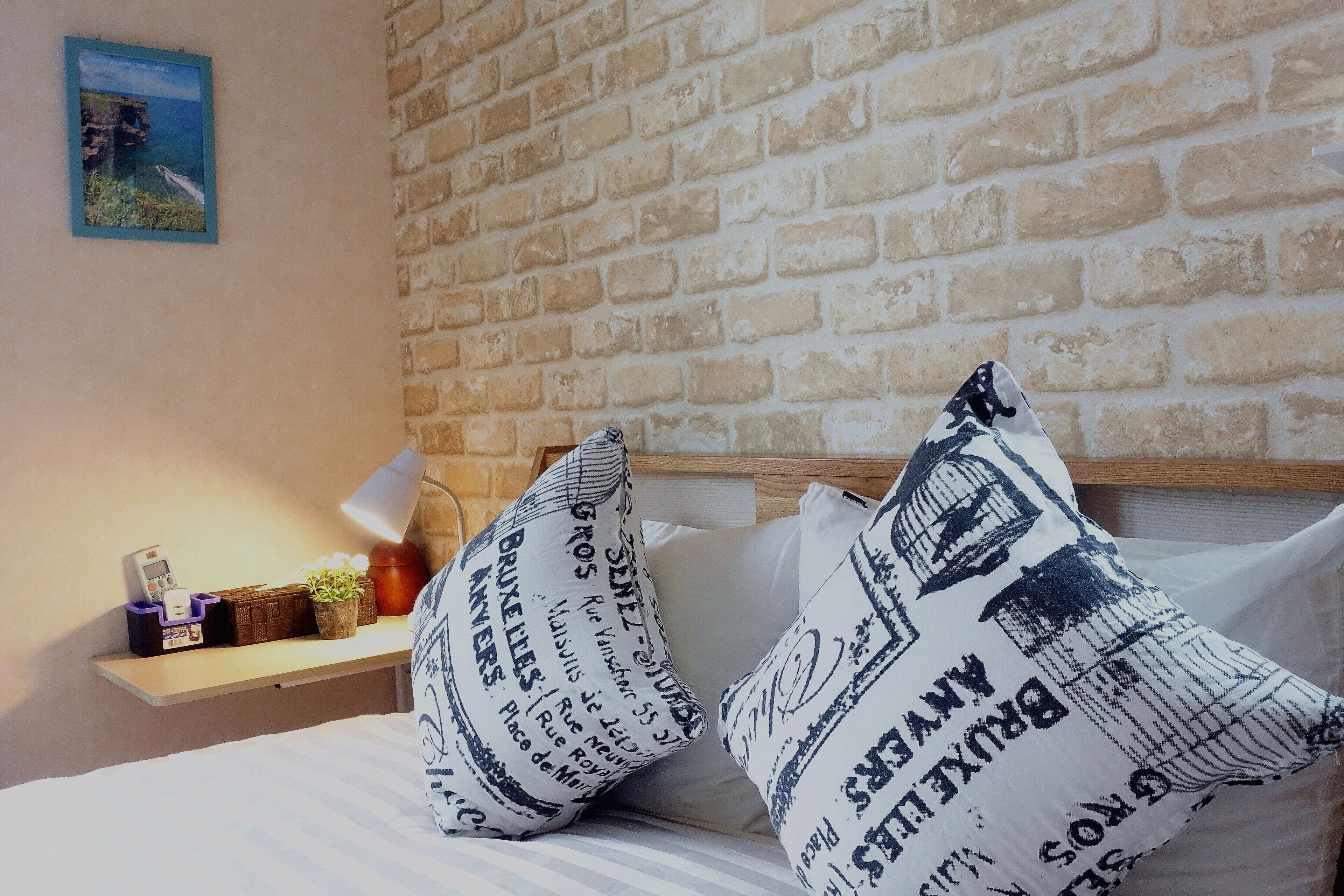 Een bed met witte lakens en twee kussens met een blauw-wit patroon, een nachtkastje met een lamp, en een bakstenen muur.