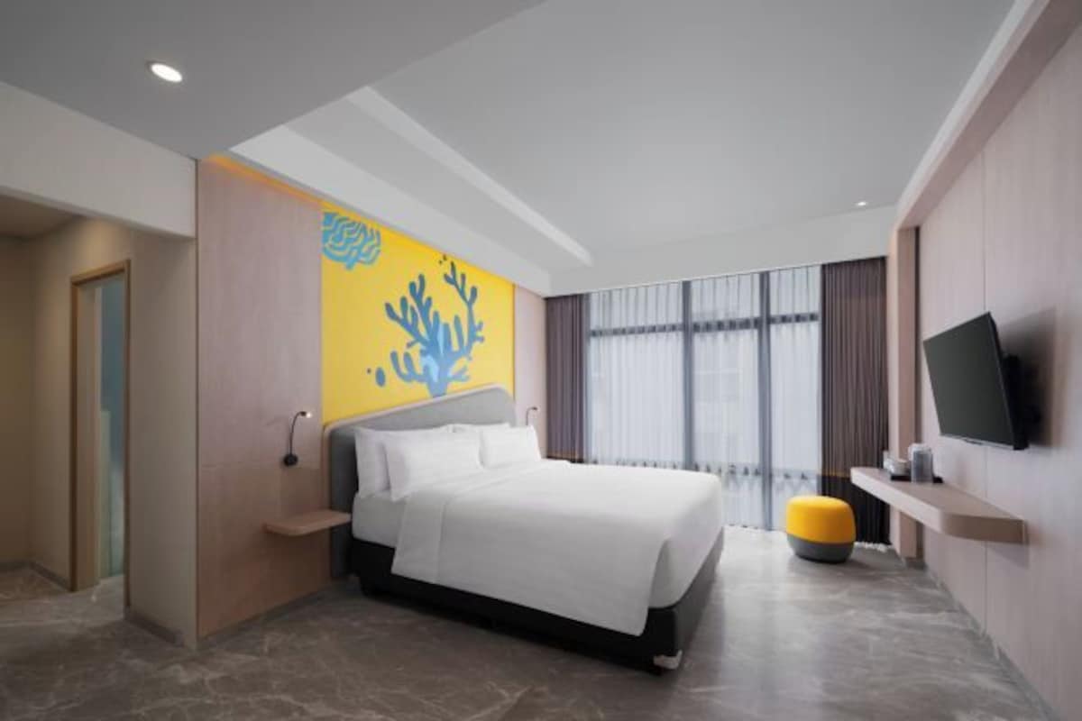 A modern hotel room with a bed, a television, a window with curtains, and a wall-mounted shelf.
