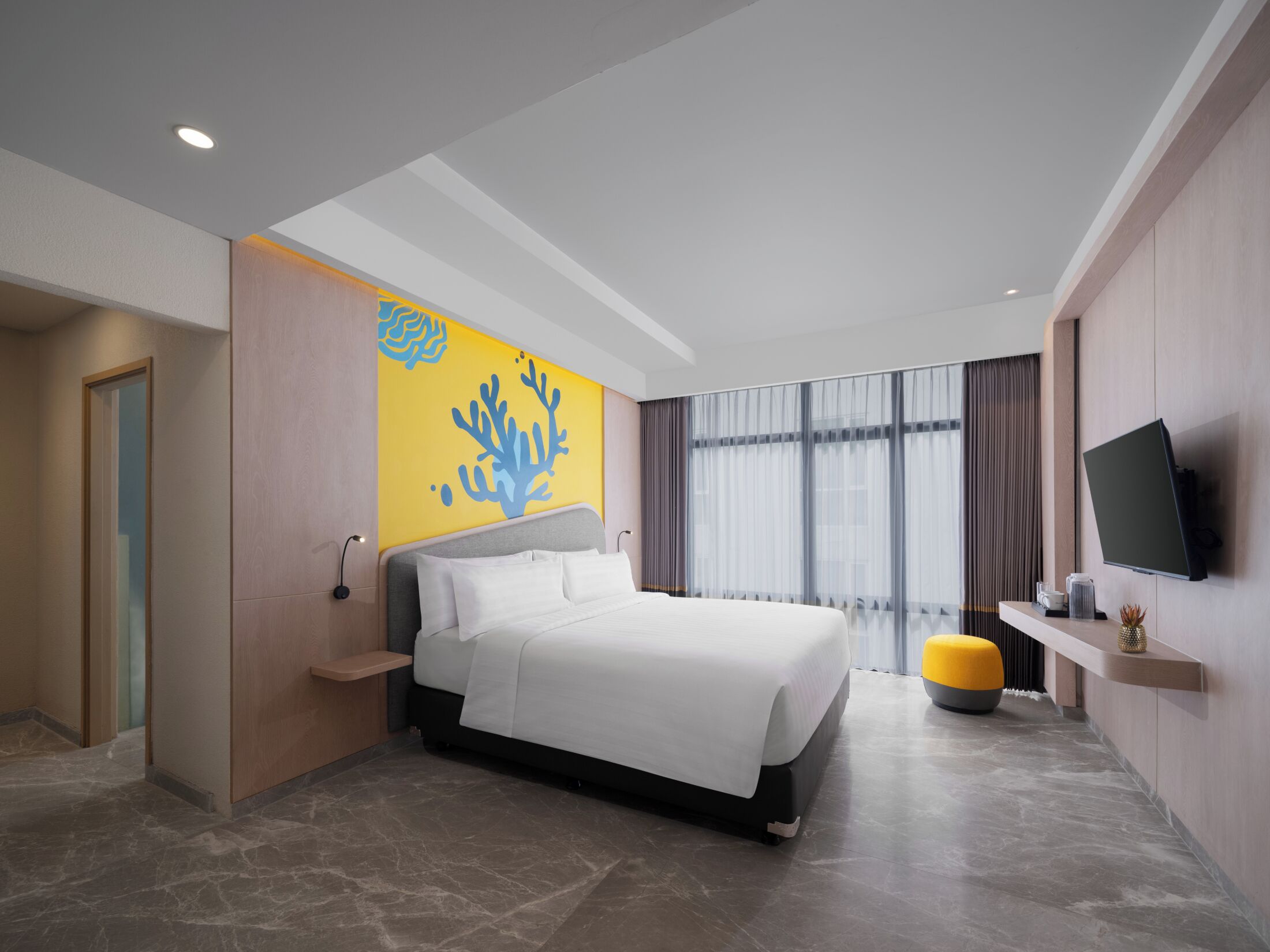 A modern hotel room with a bed, a television, a large window with curtains, and a yellow accent wall.