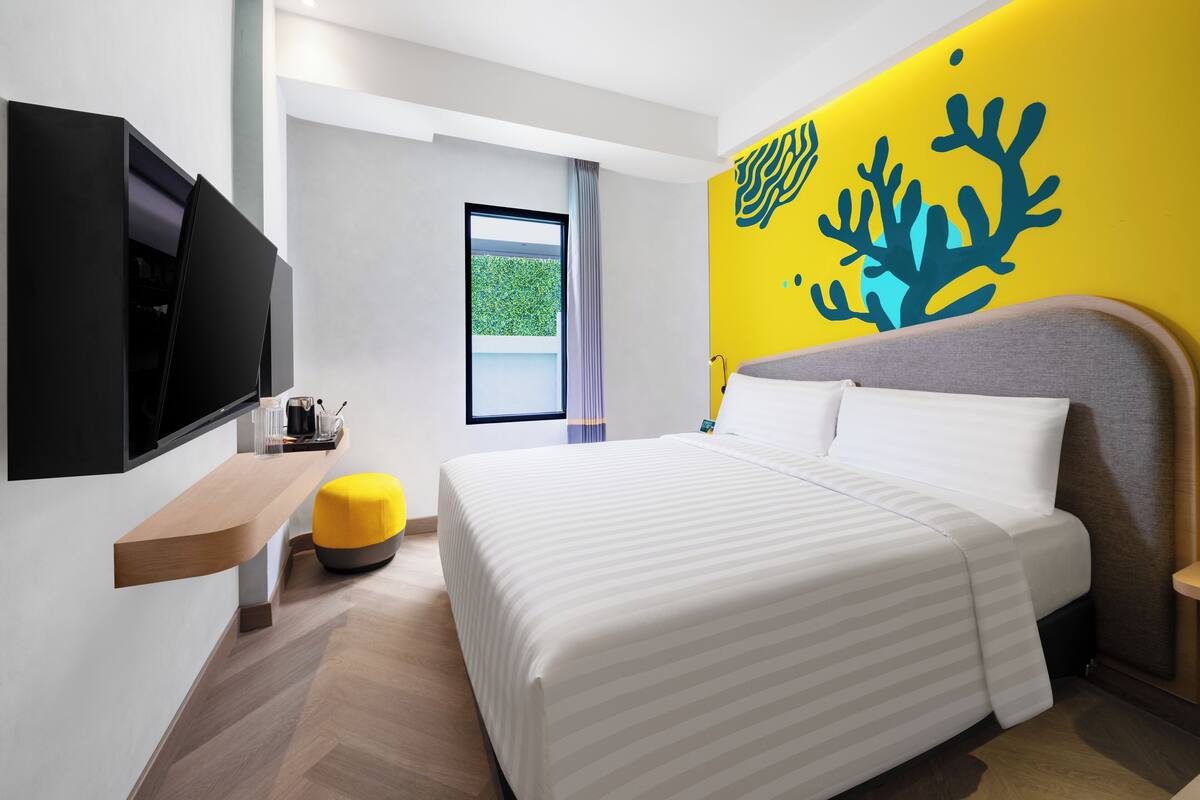 Habitació d'hotel moderna amb un llit gran, un televisor de pantalla plana fixat a la paret, una taula de fusta petita i un otomà groc.