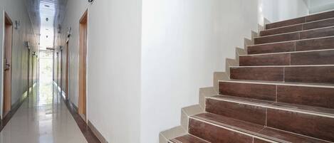 Un largo y estrecho pasillo con piso pulido y puertas de madera en un lateral, que conduce a una escalera con escalones de madera oscura y peldaños color beige.