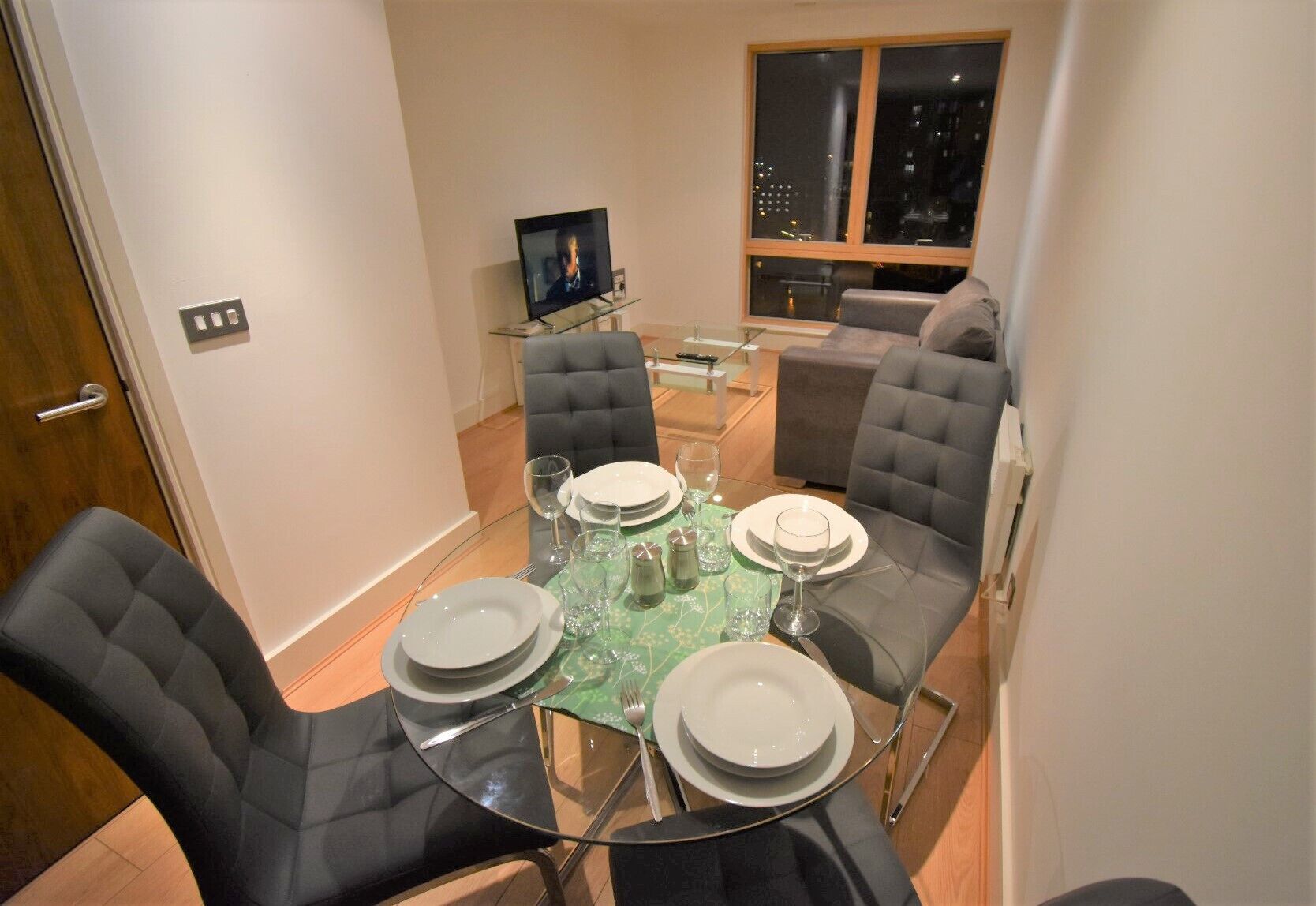 A dining area with a round table set for four, surrounded by four upholstered chairs. A flat-screen TV is mounted on the wall above the table.