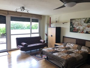 Une chambre à coucher comprenant un lit, un canapé, une armoire, un ventilateur de plafond et une grande fenêtre avec des stores.