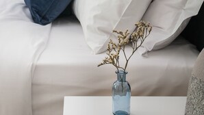 A neatly made bed with white and dark blue pillows, a white bedside table, and a blue vase with dried flowers. - Bright 2BD Apartment in Psychiko (Neo Psichiko)
