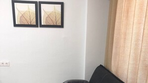 Olohuoneessa on tumma sohva, kaksi koristetyynyä, lasinen sohvapöytä ja seinällä kaksi kehystettyä taideteosta, joissa on kuvattuna lehtiä.