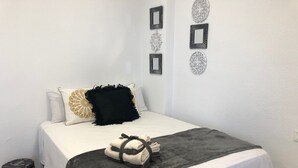 Siististi pedattu sänky, jossa on valkoinen ja harmaa peitto, musta tyyny ja koristeellinen tyyny, jossa on kultainen kuvio. Sängyn yläpuolella on seinäkoristeita, ja sängyn vieressä on pieni jakkara, jossa on musta päällinen.