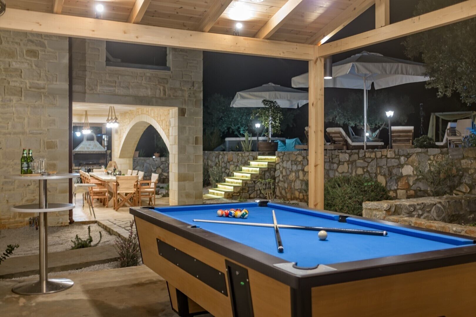 An outdoor pool area with a wooden pool table, stone walls, and a dining setup with outdoor seating.