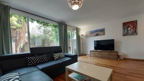 Ein modernes Wohnzimmer mit einem schwarzen Sofa, einem Couchtisch und einem Fernseher auf einer Holzkommode. Große Fenster bieten Blick auf Grünflächen.