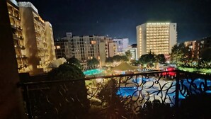 Vista noturna de um hotel com área de piscina, edifícios iluminados e uma varanda com mesa posta para um chá.