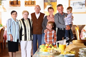Eine Familie mit sechs Personen sitzt an einem Esstisch mit Früchten und einer Kanne.
