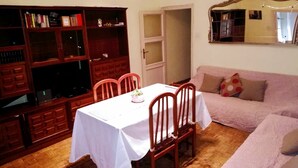 Un comedor con una mesa dispuesta para cuatro personas, rodeada de sillas de madera. A la izquierda, una estantería llena de libros y objetos decorativos. Un sofá con un cojín estampado está apoyado en la pared, y un espejo cuelga sobre él.
