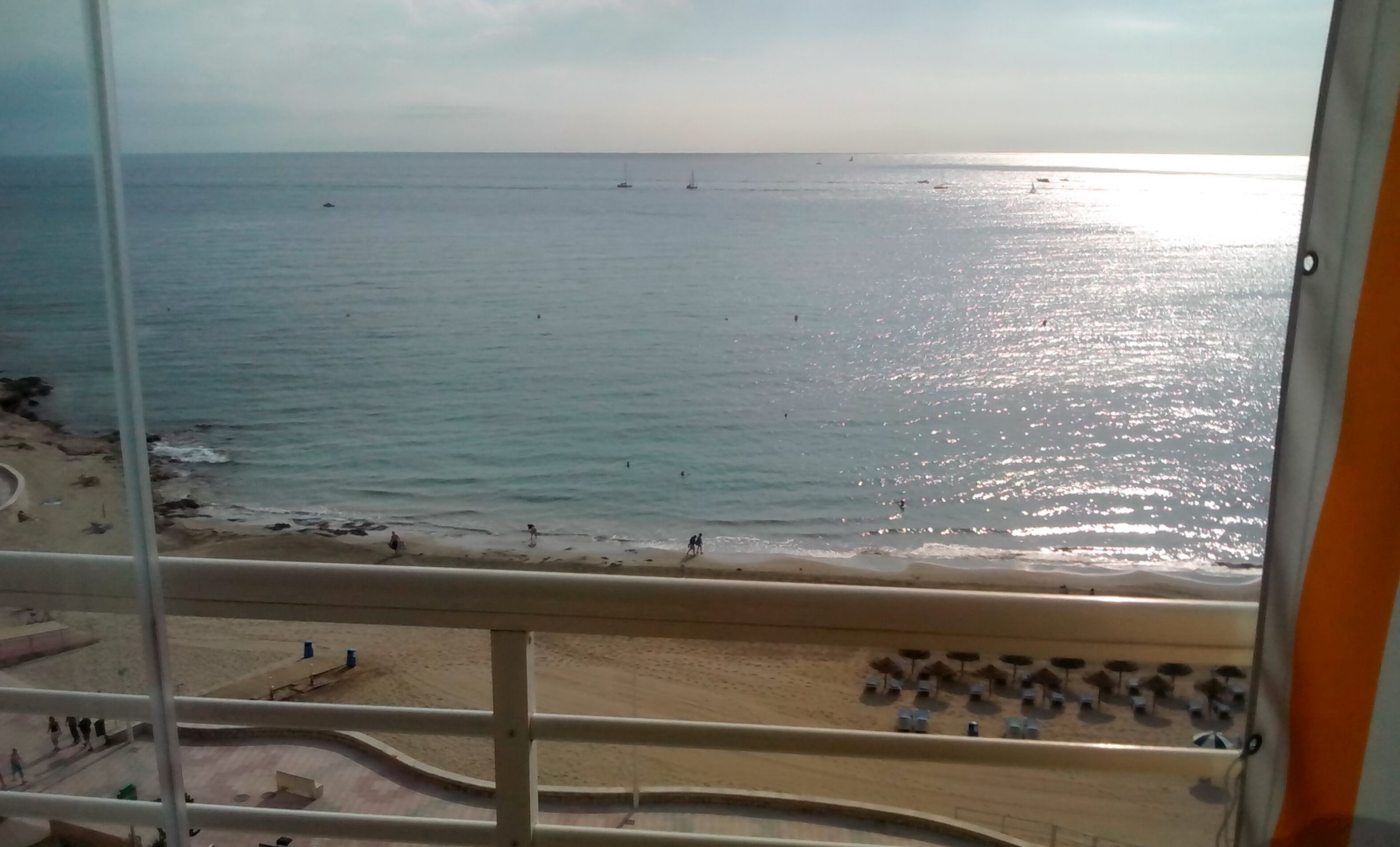 Blick aus einem Fenster auf den Strand, das Meer und eine Promenade mit Sonnenschirmen und Sitzgelegenheiten.