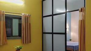 A room with yellow walls, a ceiling fan, a bed with a blue checkered bedspread, and a small table with a plant and papers. - RP Studio Rooms (Igatpuri)