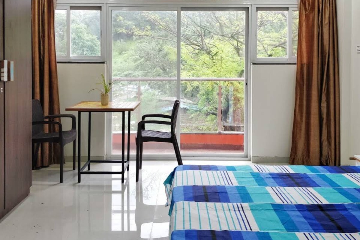 A bedroom with a bed, a desk, a chair, and a view of greenery through large windows.