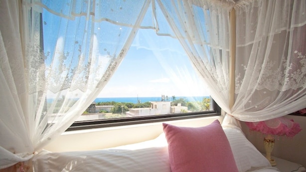 Ein Schlafzimmer mit einem Bett, einem rosa Kissen und Blick auf Gebäude durch leichte Vorhänge.