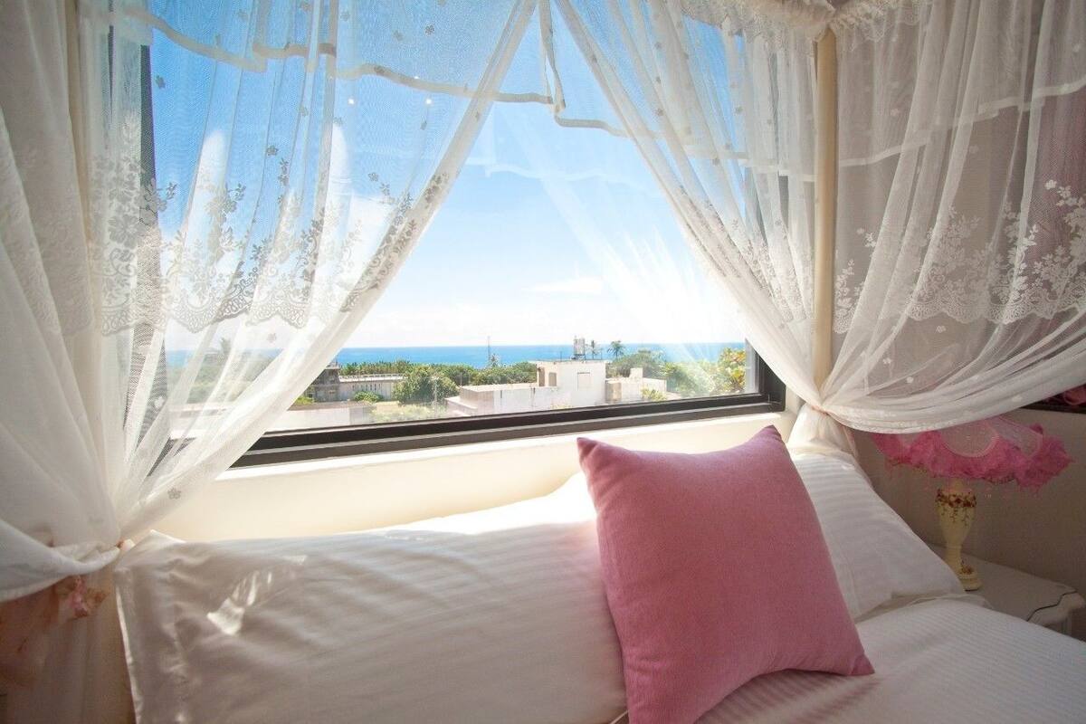 A bedroom with a bed, a pink pillow, and a view of buildings through sheer curtains.