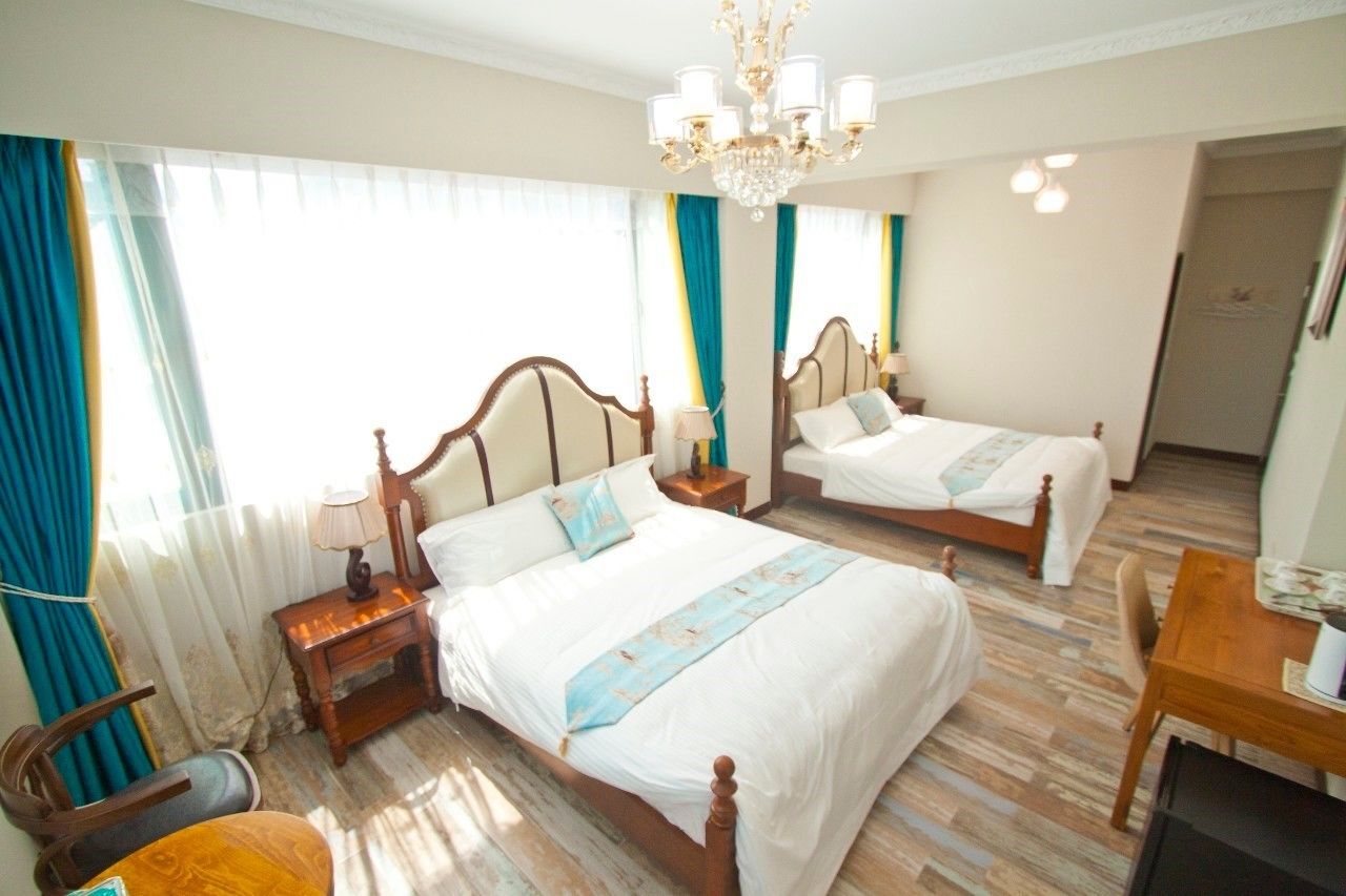 A hotel room with two single beds, a chandelier, a wooden nightstand with a lamp, a wooden chair, and a small table.