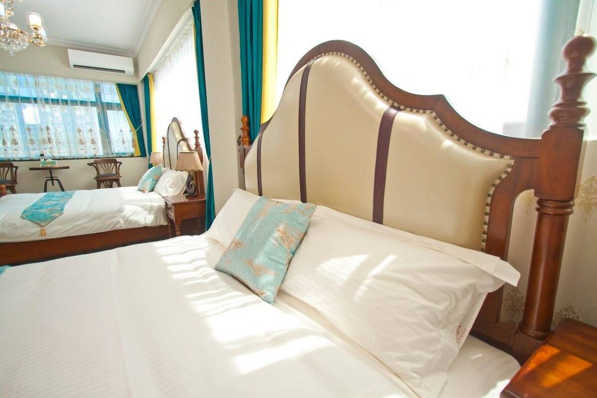 A hotel room with two beds, a chandelier, a window with curtains, and a small table with a lamp.