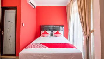 A hotel room with a red wall, a bed with white and red bedding, a window with beige curtains, and a door with a frosted glass panel.