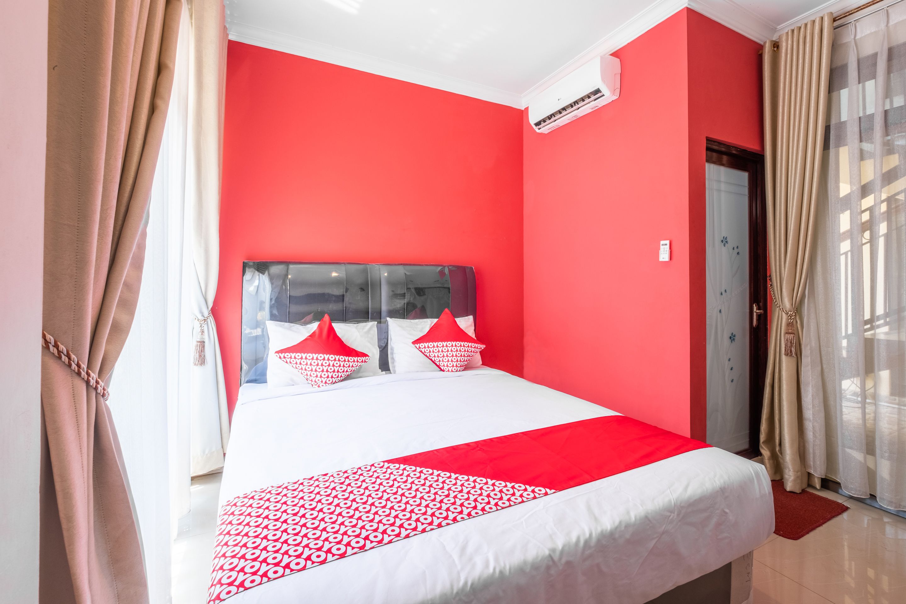 Une chambre d’hôtel avec un mur rouge, un lit recouvert d’une courtepointe à motifs rouges et blancs, et une fenêtre ornée de rideaux beiges.