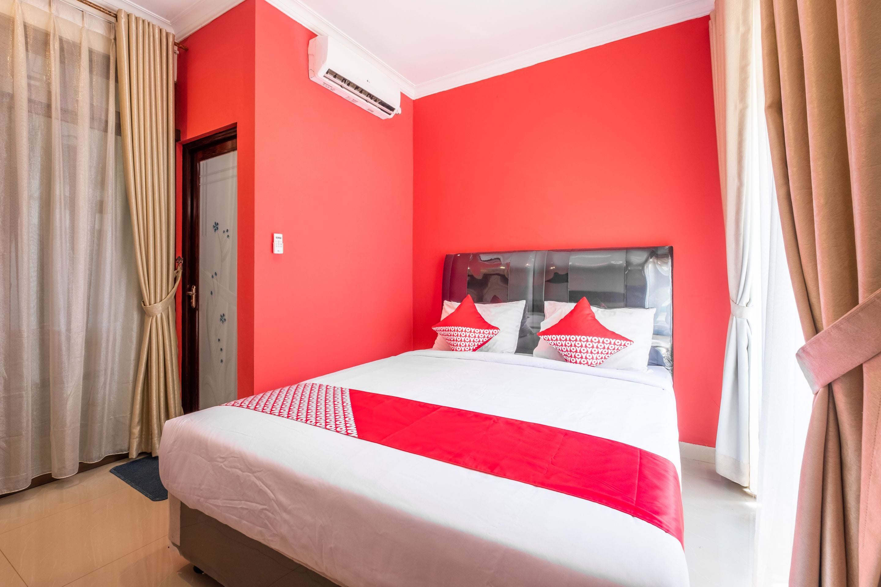 A hotel room with a red wall, a bed with white and red bedding, beige curtains, and an air conditioning unit.