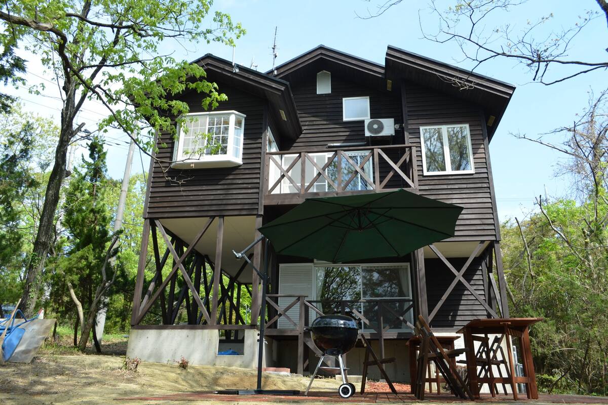 A two-story wooden house with a balcony, a grill, and an umbrella.