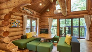 A log cabin interior with a wooden dining table, green sofas, and large windows offering a view of the outdoors. - Family Cottage NOTOIE (Shika)