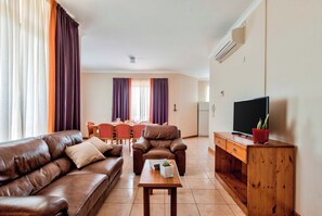 A living room with a brown leather sofa, a wooden coffee table, a TV on a wooden stand, and a dining area with a table and chairs.